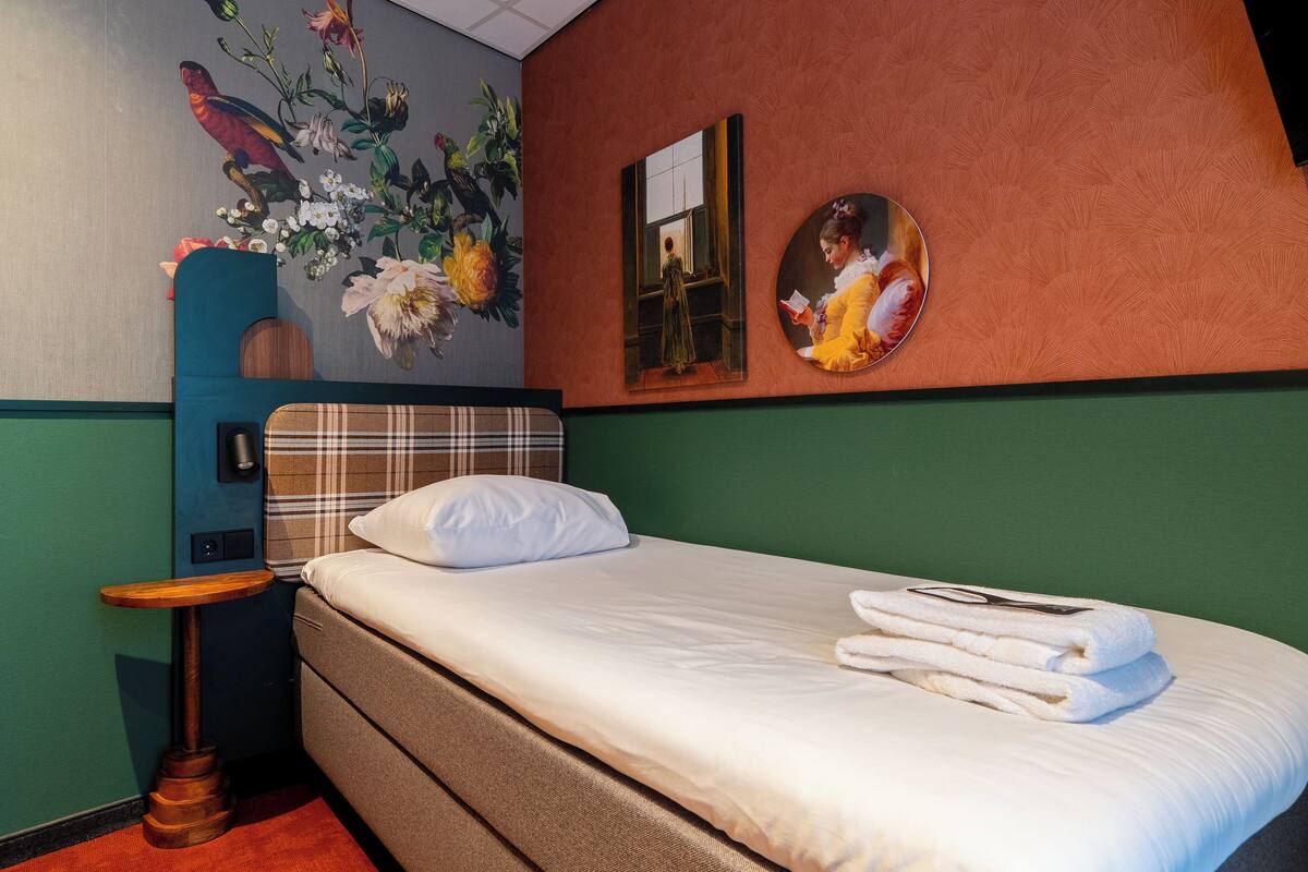 A hotel room with a bed, a nightstand, a wall-mounted lamp, a painting, and a mirror.