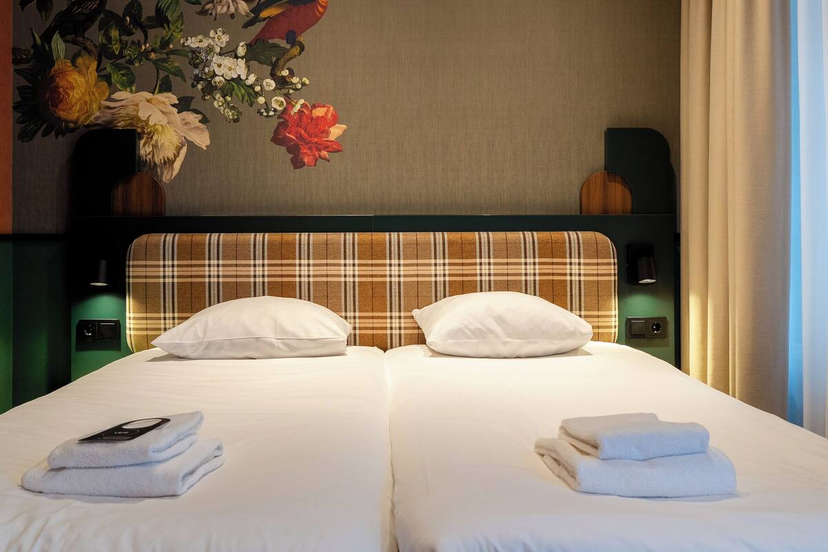 A hotel room with two beds, a large floral artwork above the beds, and a view of the outdoors through a window.