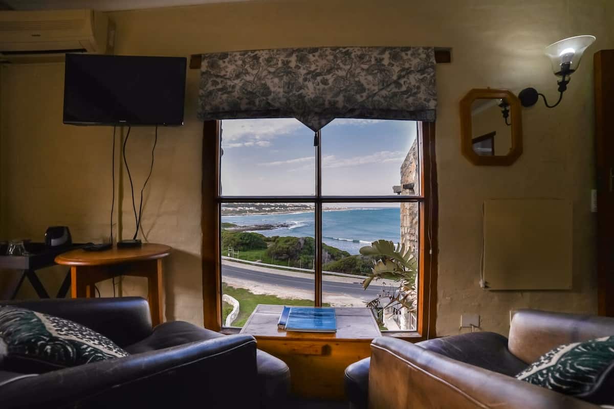 A living room with a large window offering a view of the sea and a road. There is a television on the wall, a small table, and two sofas.