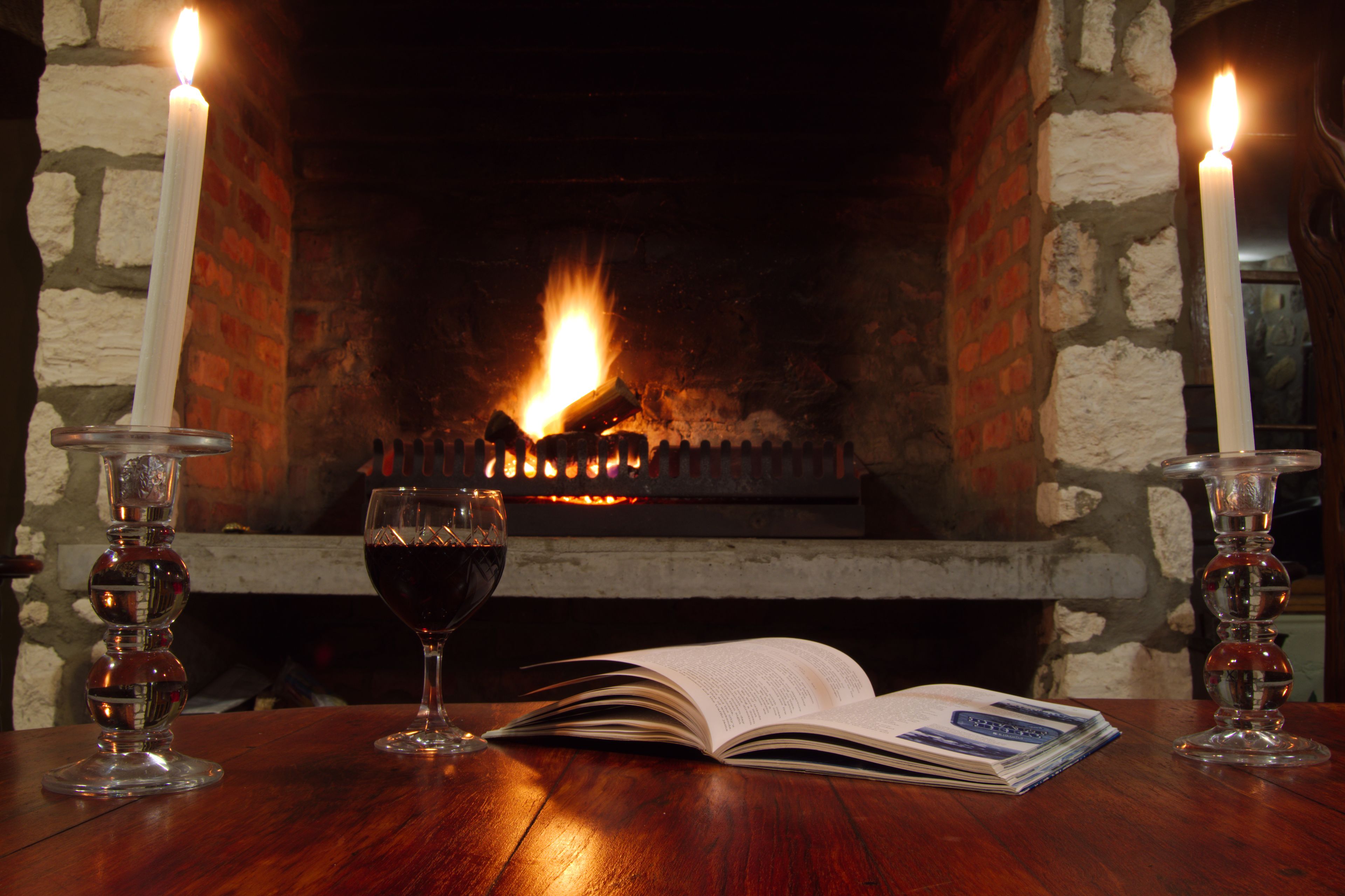 A fireplace with two candles, a glass of red wine, and an open book on a wooden table.