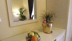 A room with a white counter, a bowl of fruit, a plant in a gold pot, and a mirror reflecting a plant and a window with curtains. - Il Monte (Monte San Savino)