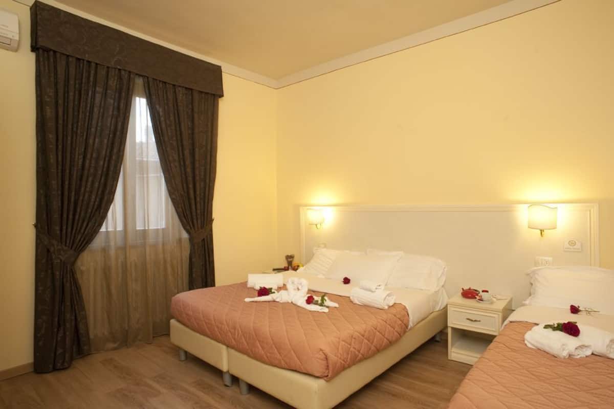 A hotel room with two beds, a large window with curtains, bedside lamps, and a bouquet of flowers on the beds.