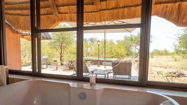 A bathroom with a large bathtub, a window with a view of outdoor furniture and trees. - Mohlabetsi Safari Lodge (Hoedspruit)