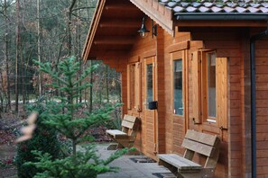 A wooden cabin with a covered porch, benches, and a small tree in the foreground.