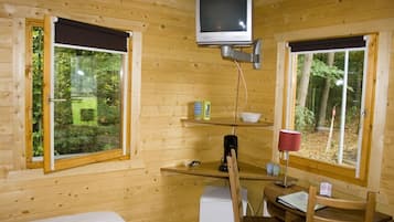 Een houten kamer met een bed, een bureau met stoel, een aan de muur bevestigde televisie en ramen met uitzicht op een bos.