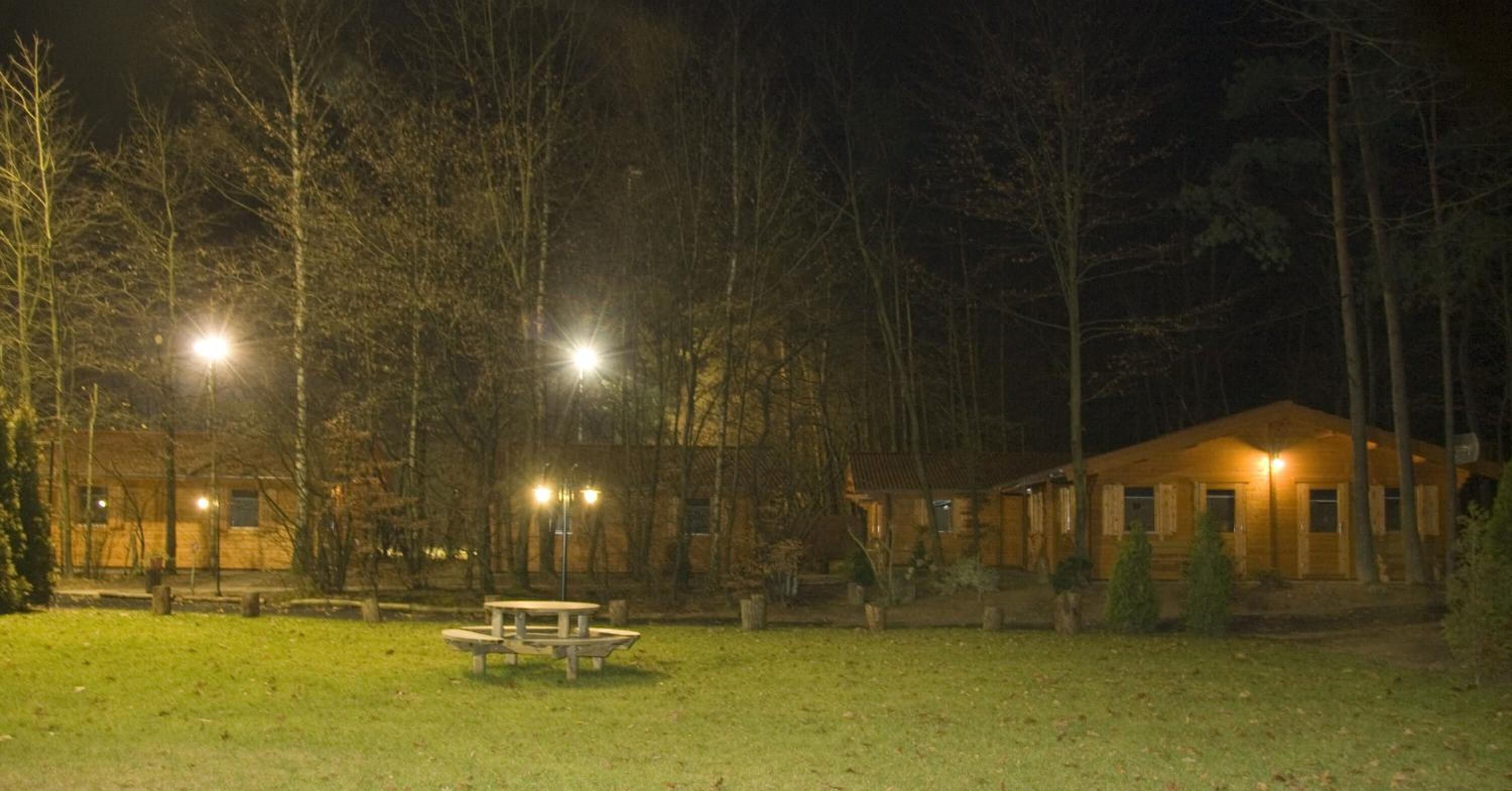 Une scène nocturne représentant un chalet avec une table de pique-nique au premier plan, entouré d’arbres et d’un bâtiment bien éclairé.