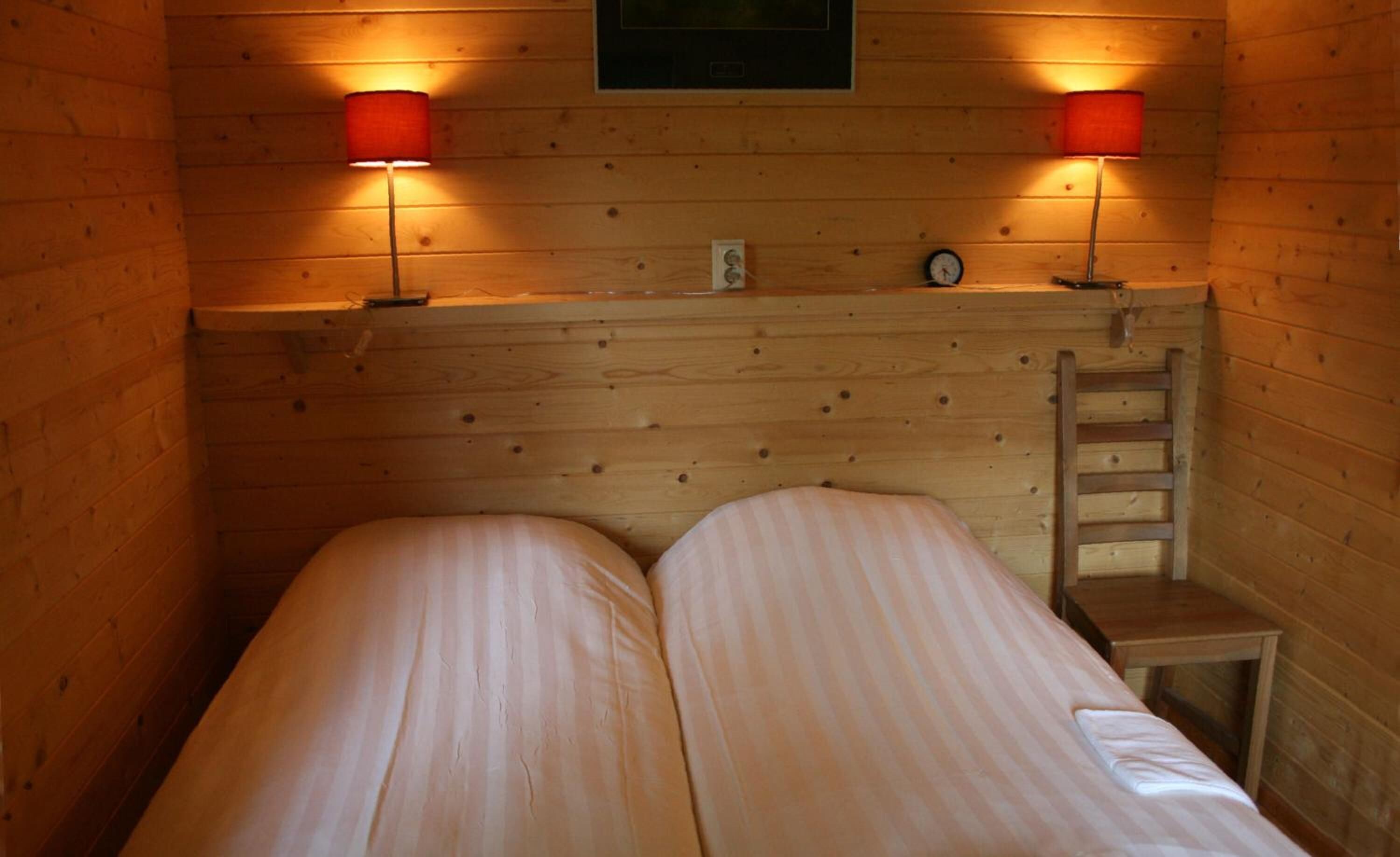 Une chambre dans un chalet en bois, avec deux lits, deux lampes rouges, une horloge et une échelle.