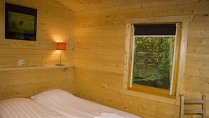 A wooden cabin room with a bed, a window with a view of trees, and a wall-mounted lamp. -  Lodges Veluwse Poort (Dieren)
