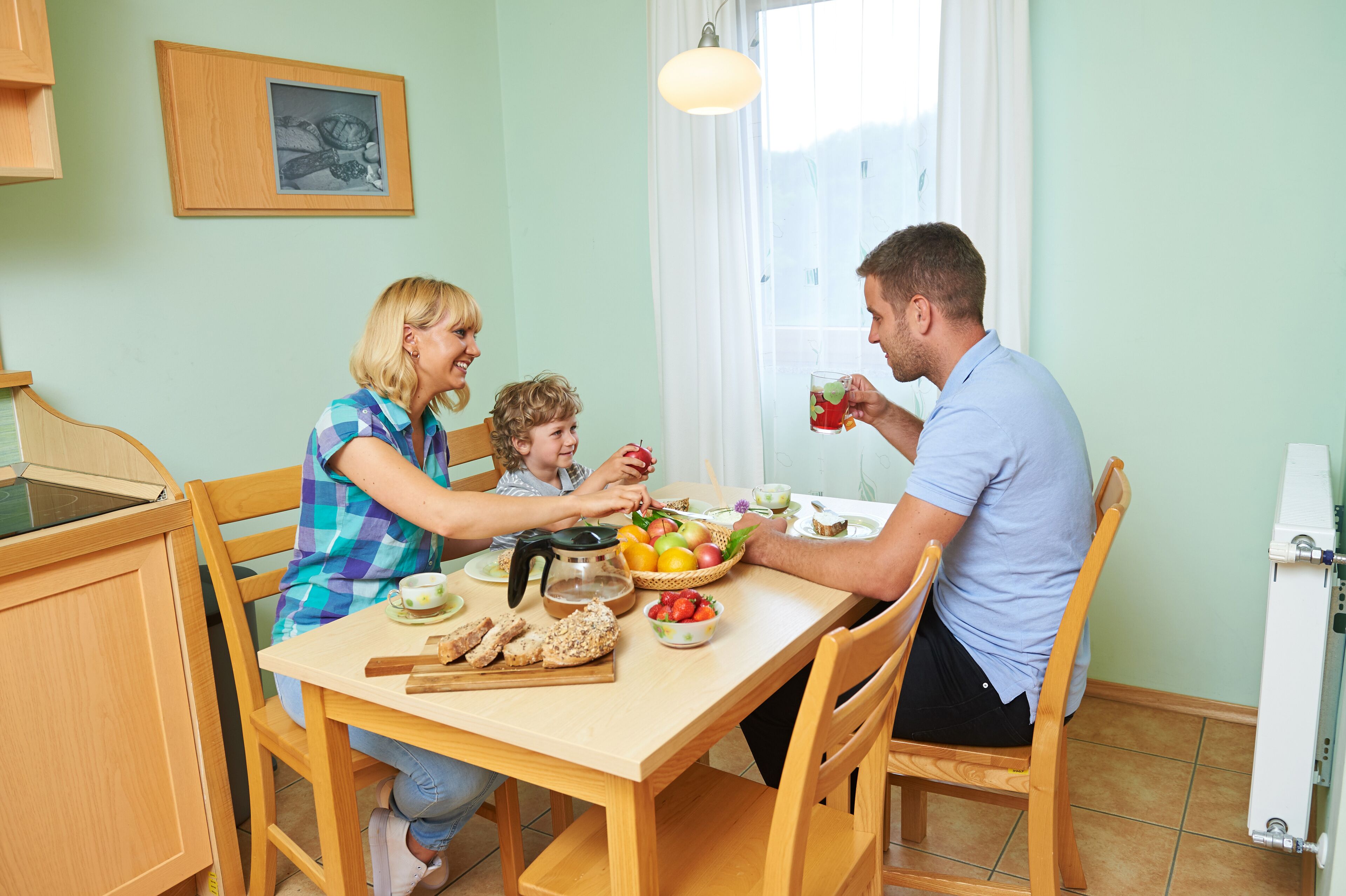 Eine Familie isst an einem Holztisch, auf dem sich eine Fruchtschale, ein Teekessel und eine Tasse befinden.