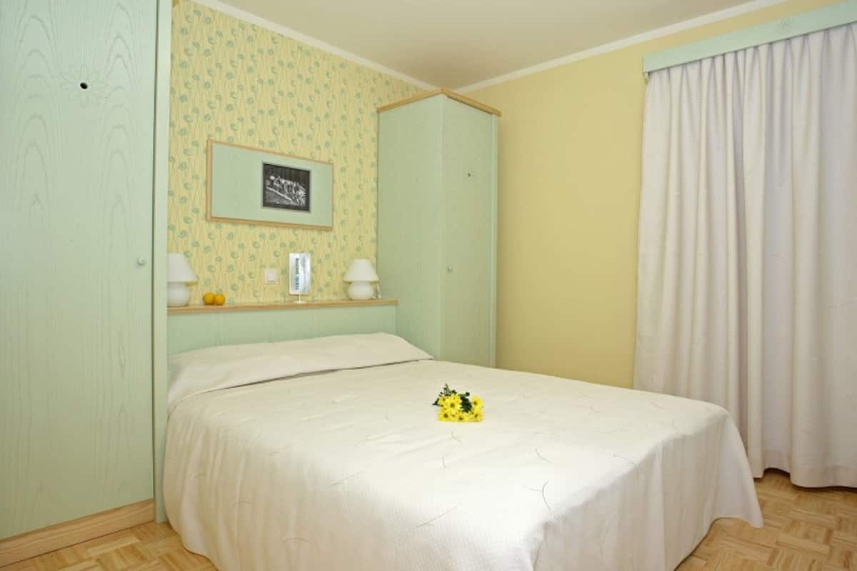 A bedroom with a bed, a nightstand, a mirror, a wall-mounted picture, and a window with curtains.