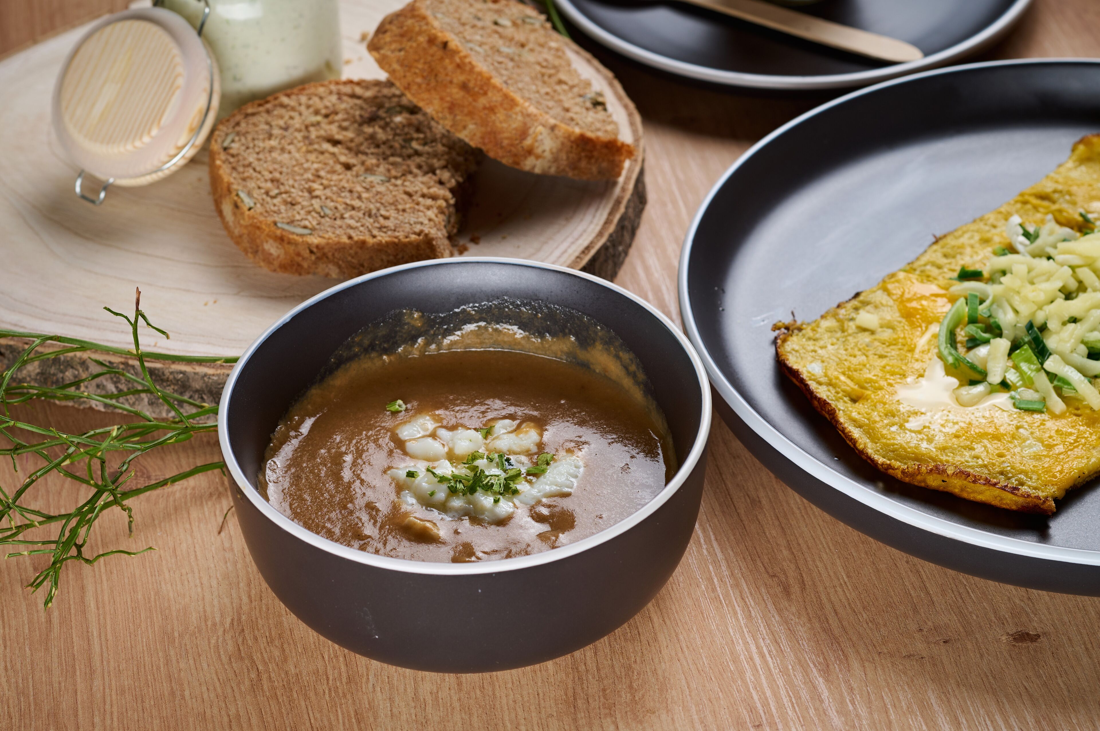 Una mesa de madera con un tazón de sopa, rebanadas de pan y un plato con un huevo frito y cebollines.