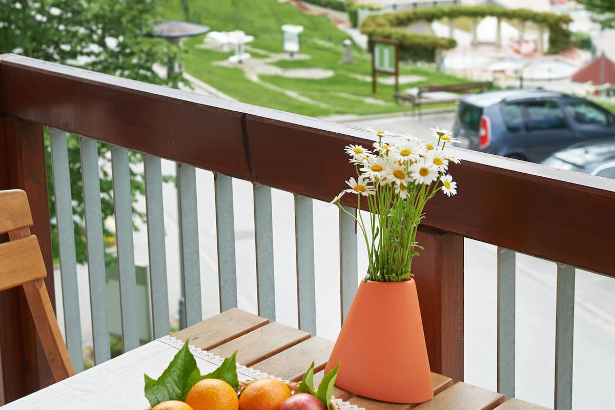 A balcony with a table set with a fruit bowl and a vase of flowers, overlooking a park with a playground and a building in the background.