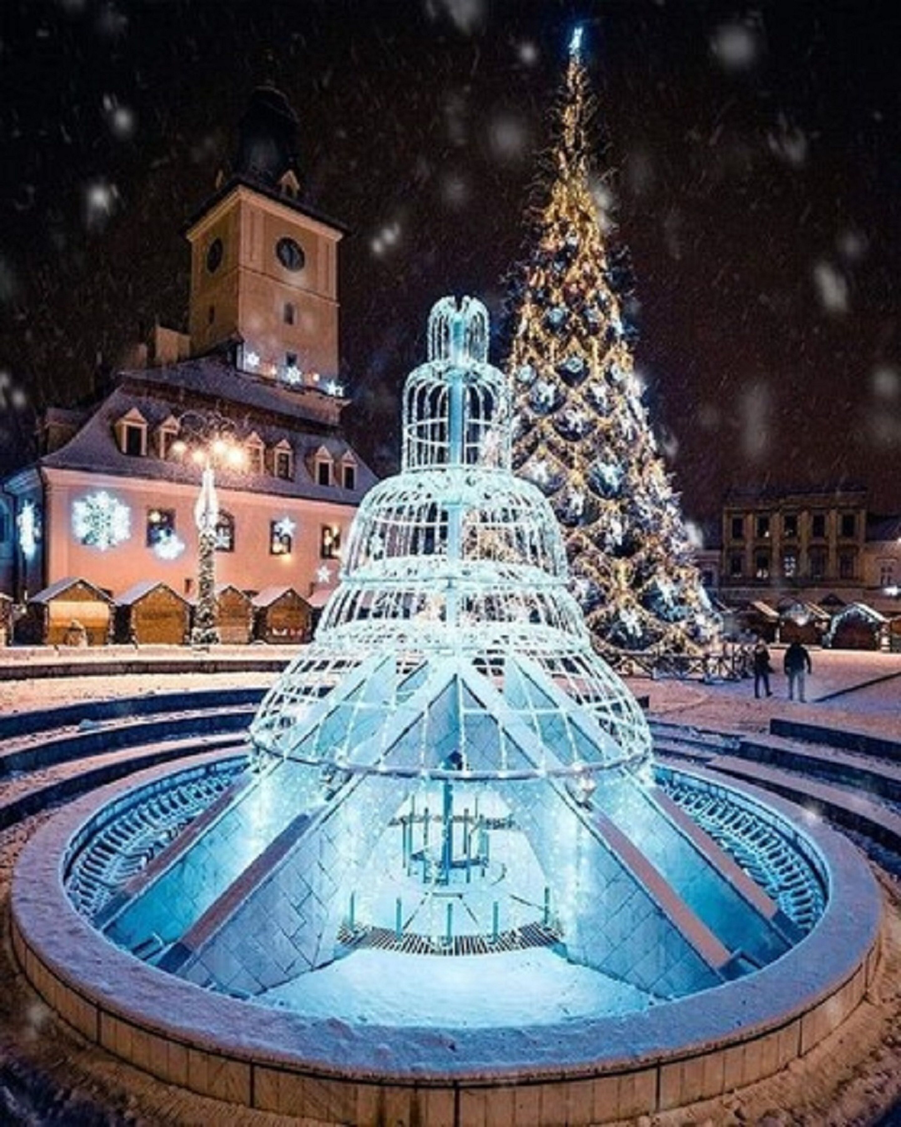 A large, illuminated water fountain with a geometric structure in the center, surrounded by a circular brick platform.