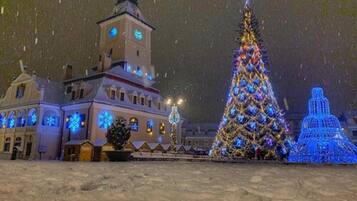 A snow-covered plaza with a large Christmas tree and a building with illuminated windows and a clock tower.