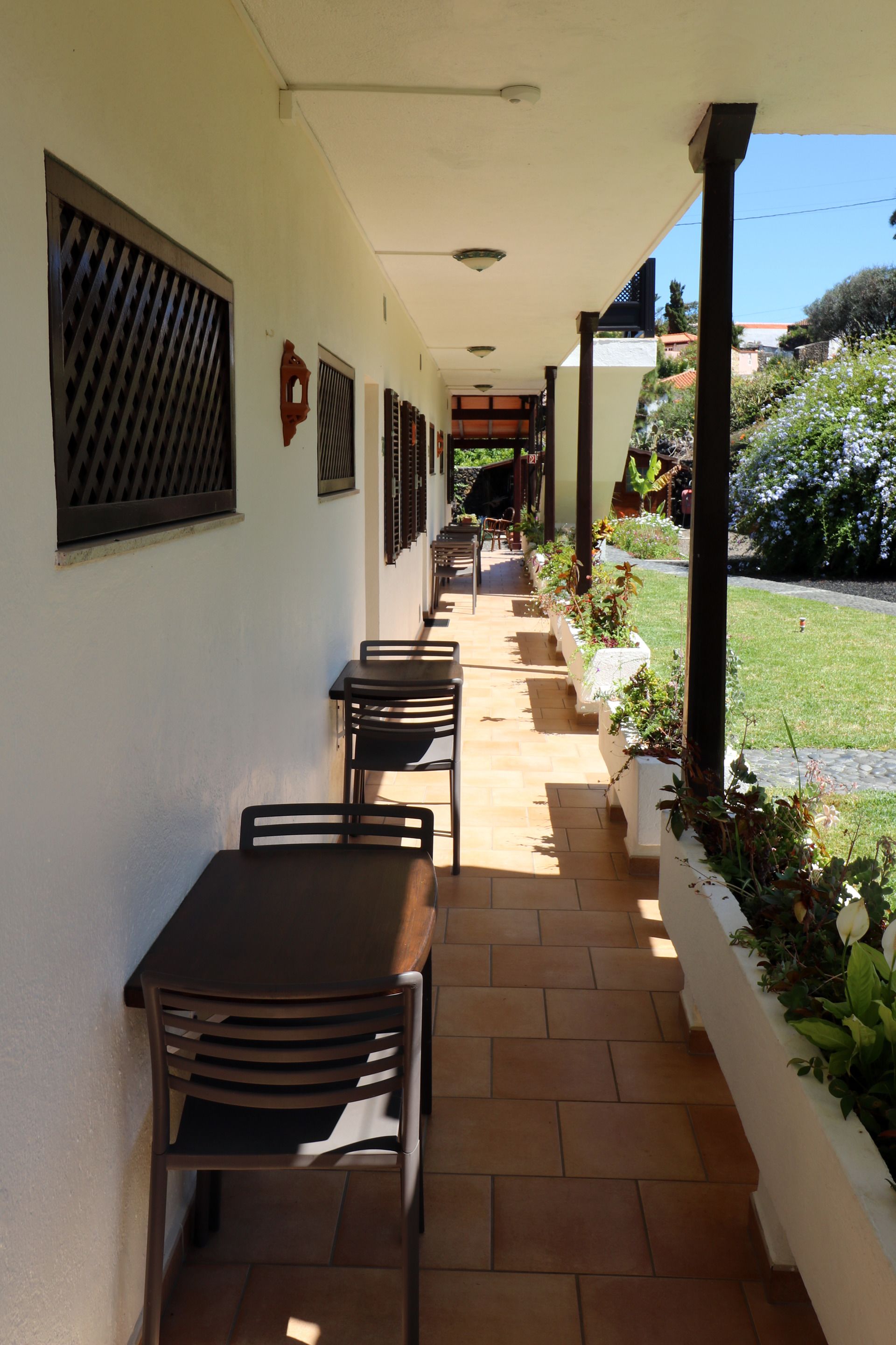 Un espacio al aire libre cubierto con varias mesas y sillas, piso de baldosas y un jardín con plantas y flores.