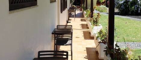 Un espacio al aire libre cubierto con varias mesas y sillas, piso de baldosas y un jardín con plantas y flores.