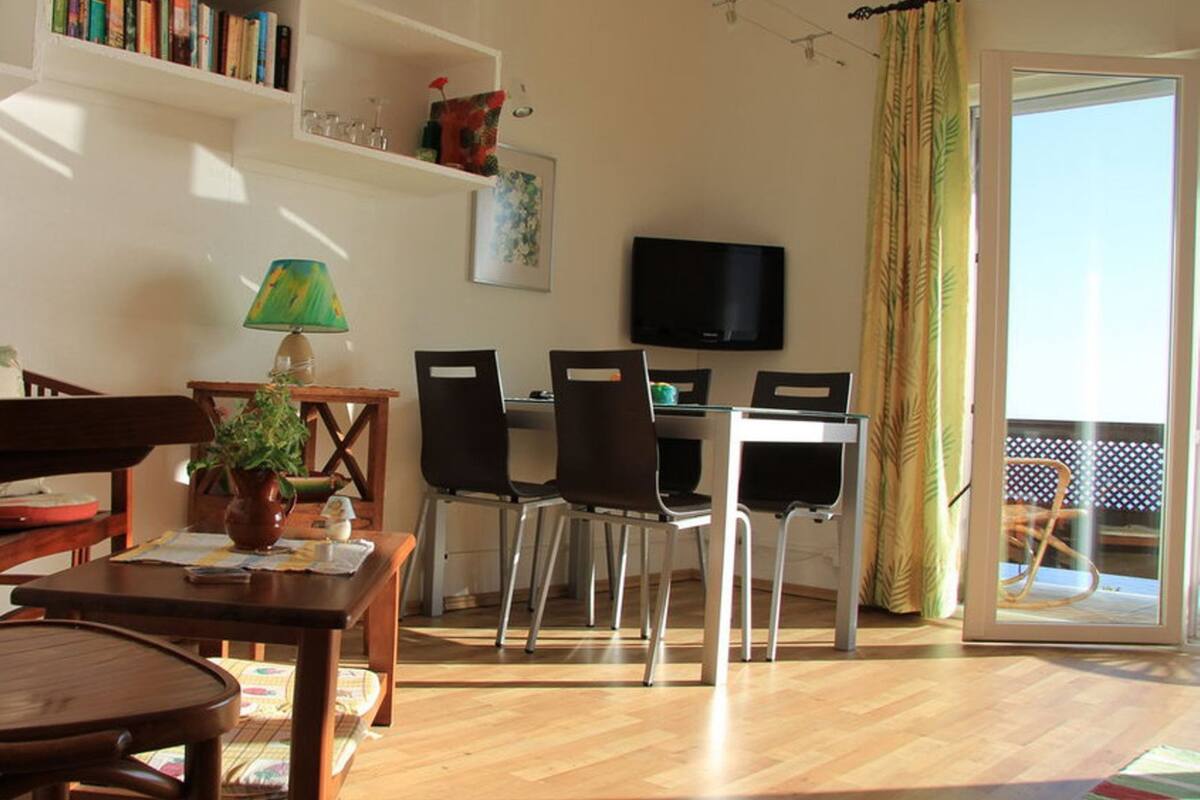 Sala de estar con una mesa de madera, sillas, televisor y un balcón con cortina amarilla.