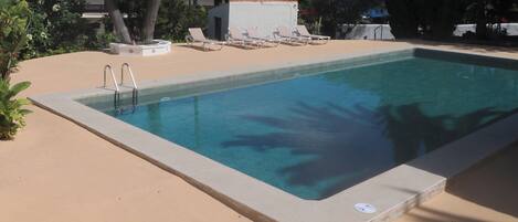 Una piscina rectangular con borde de hormigón, rodeada por una zona arenosa con sillas de descanso y una pequeña estructura.
