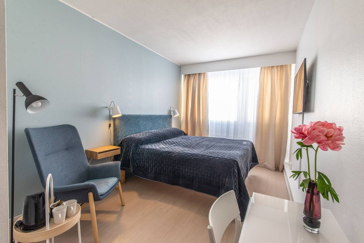 A hotel room with a bed, a chair, a desk, a lamp, a vase with a flower, and a TV on the wall.