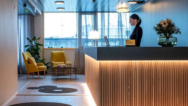 A modern reception area with a wooden reception desk, two yellow armchairs, a large window with a view of the city, and a vase with flowers on the desk.