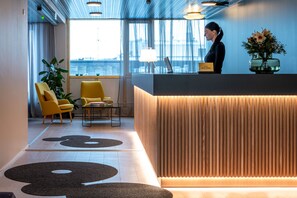 A modern reception area with a wooden reception desk, two yellow armchairs, a large window with a view of the city, and a vase with flowers on the desk. - Hotel Sparre (Porvoo)