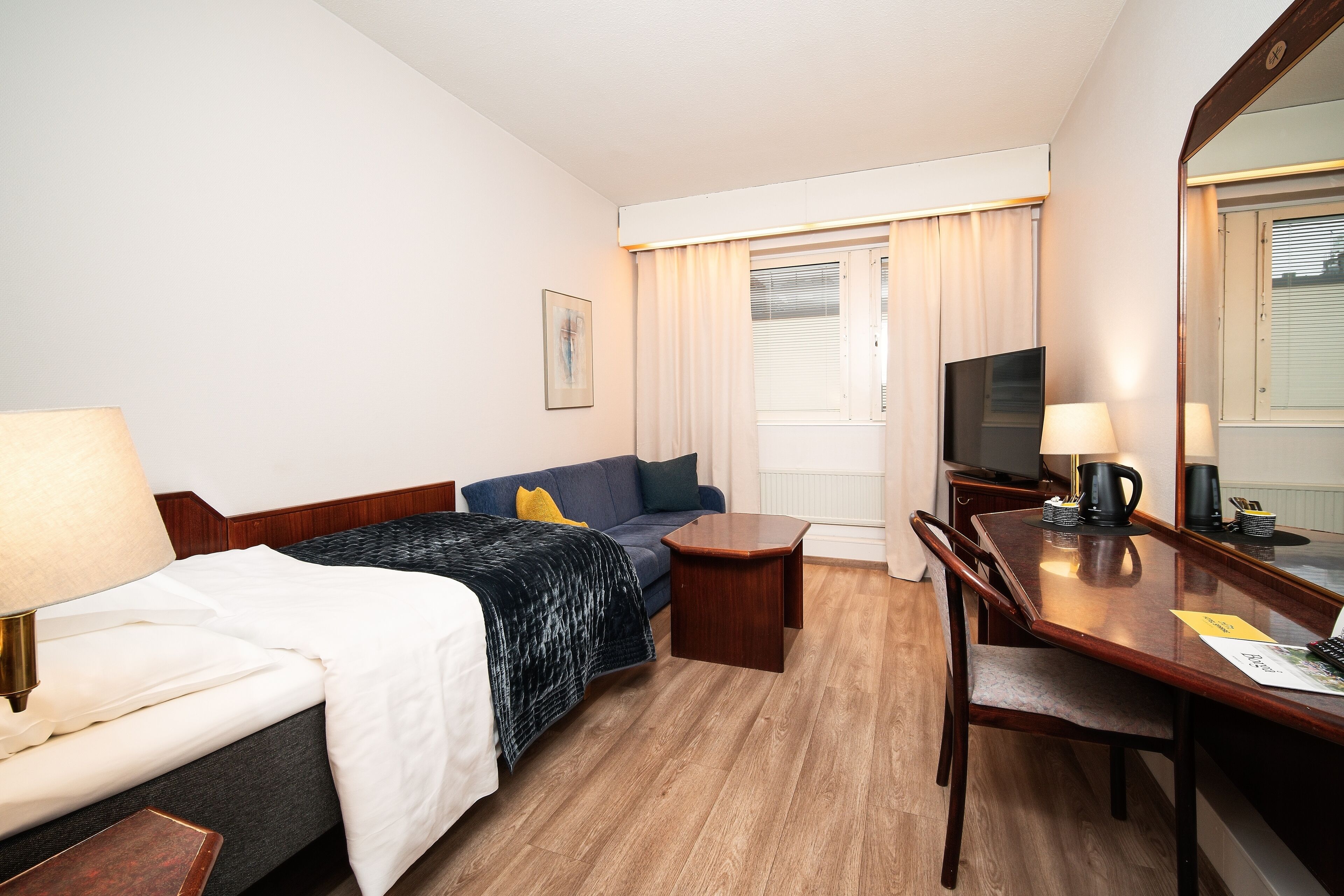 Habitación de hotel con cama, sofá, escritorio, silla, televisión y ventana con cortinas.