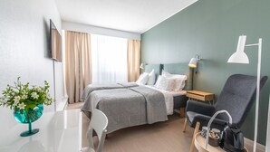 A hotel room with two beds, a desk, a chair, a small table, a lamp, and a wall-mounted TV.