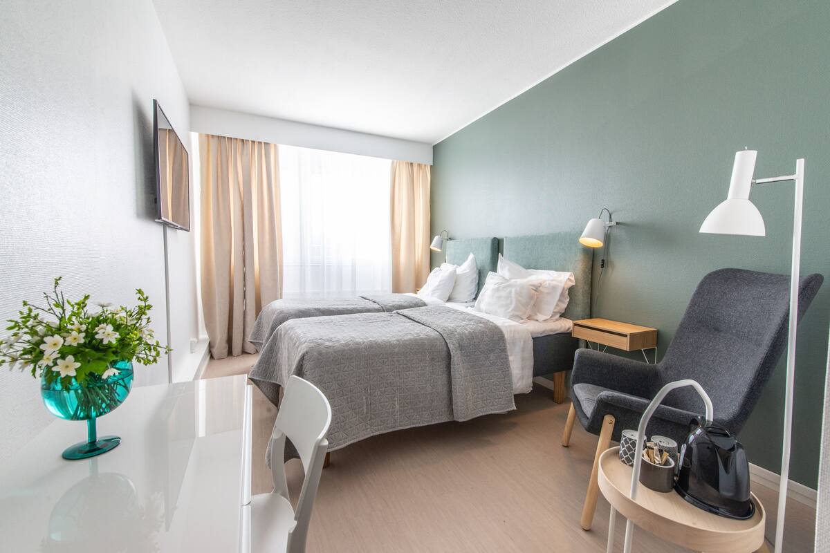 A hotel room with two beds, a desk, a chair, a small table, a lamp, and a wall-mounted TV.