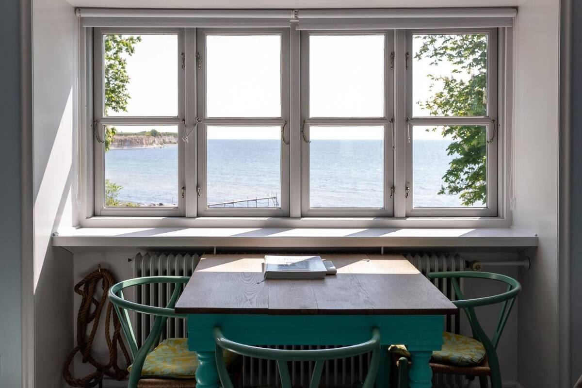 A room with a table and chairs, a view of the sea through a large window, and a radiator below the window.