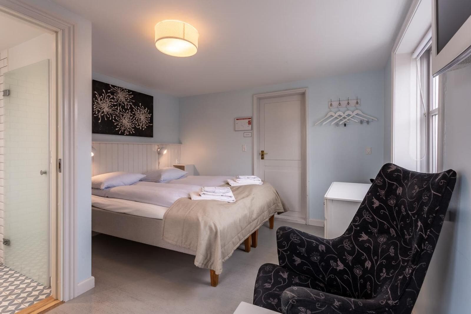 A bedroom with two beds, a chair, a bedside table, and a wall-mounted light fixture.