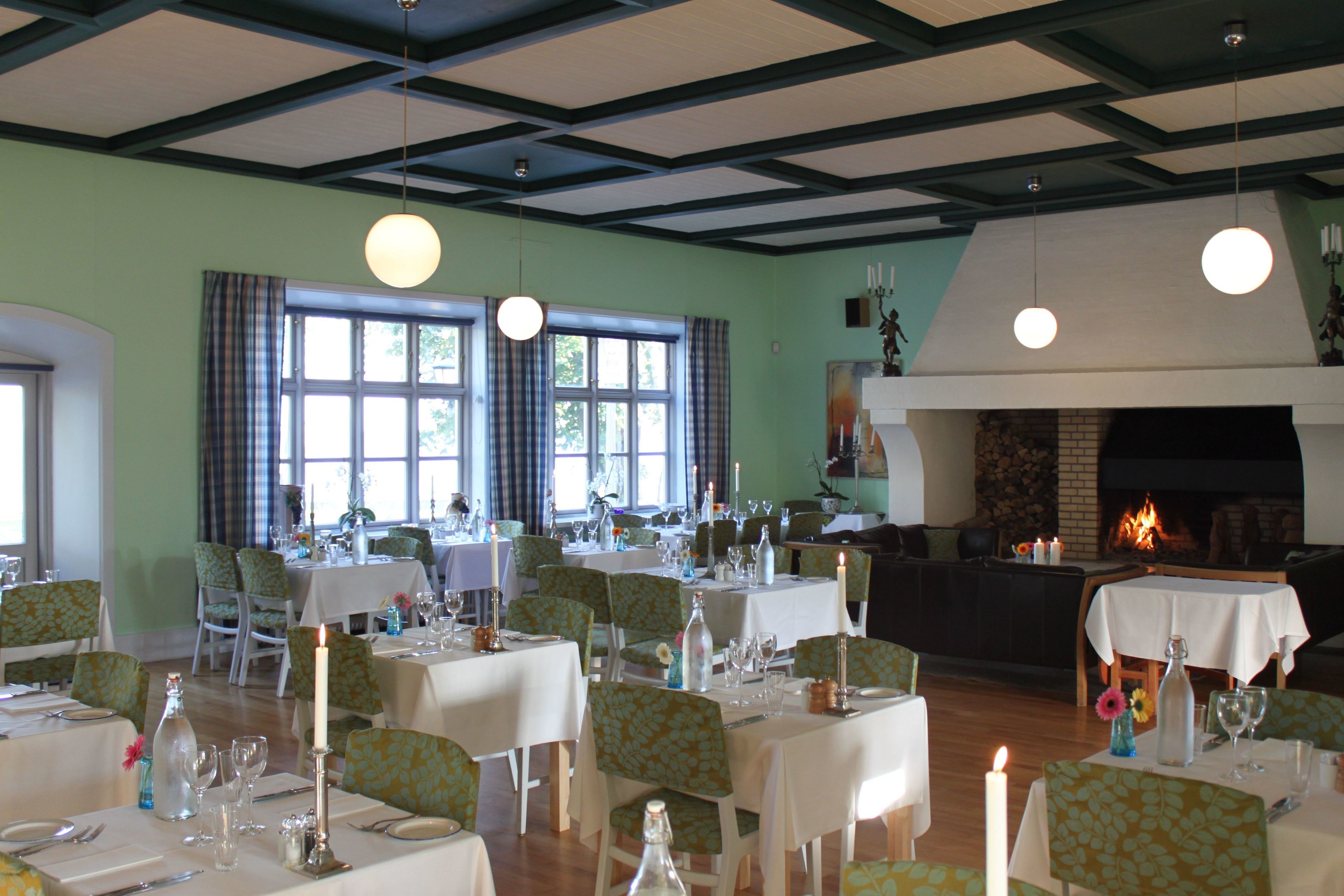 A dining area with multiple tables set for guests, a fireplace, and large windows allowing natural light.