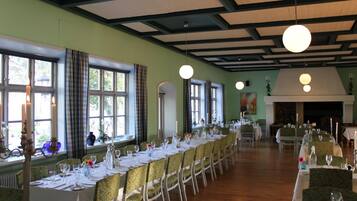 A long dining table set with white tablecloths and chairs, large windows with curtains, and a high ceiling with exposed beams.