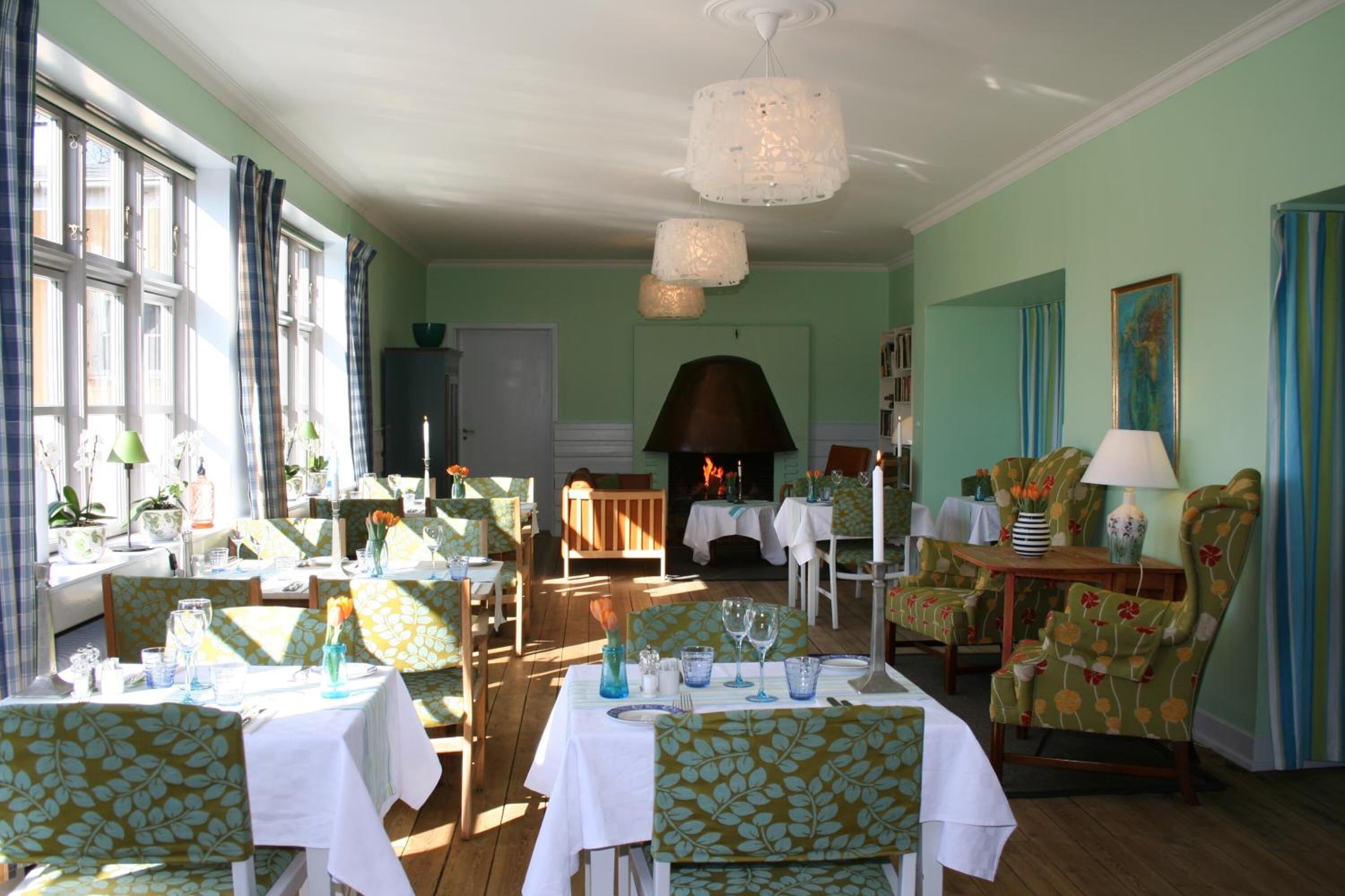 Uma sala de jantar com mesas postas para uma refeição, cadeiras com estofado floral e uma lareira ao fundo.
