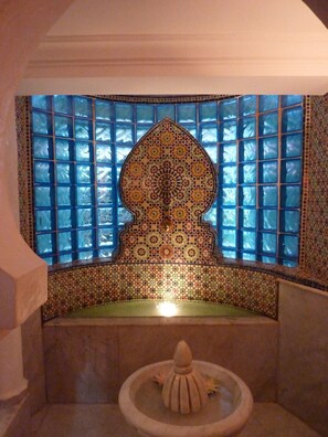 Een badkamer met een nis betegeld met mozaïek, een stenen wasbak en een raam met blauw getinte glas.