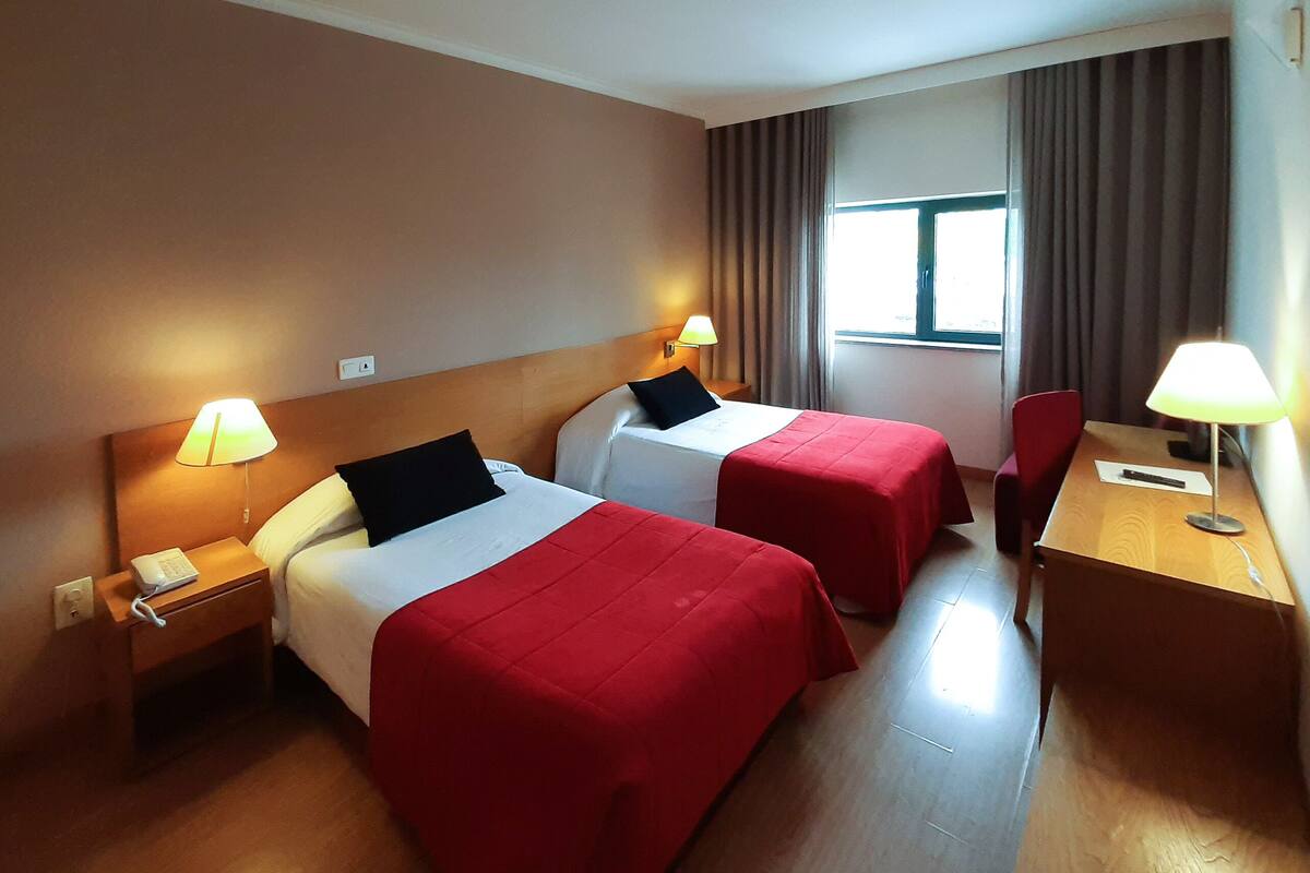 A hotel room with two beds, each with a red bedspread and black pillows, a wooden desk with a lamp, and a chair.