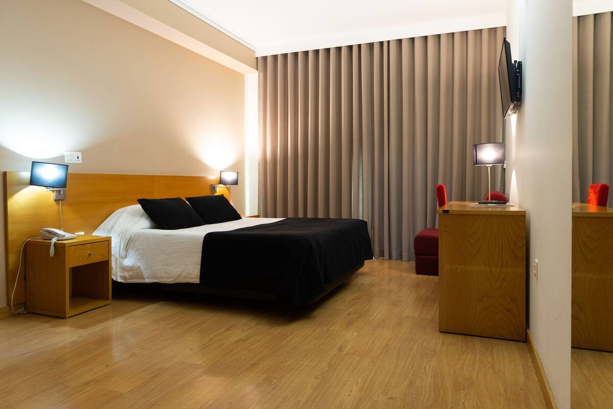 A hotel room with a wooden headboard bed, a bedside table with a lamp, a desk with a chair, and a TV mounted on the wall.