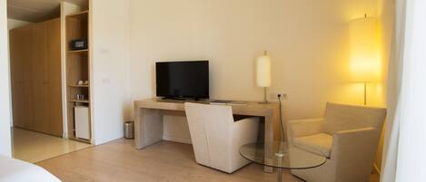 A modern hotel room with a wooden desk, a television, a glass table, a beige armchair, and a bedside table with a lamp.