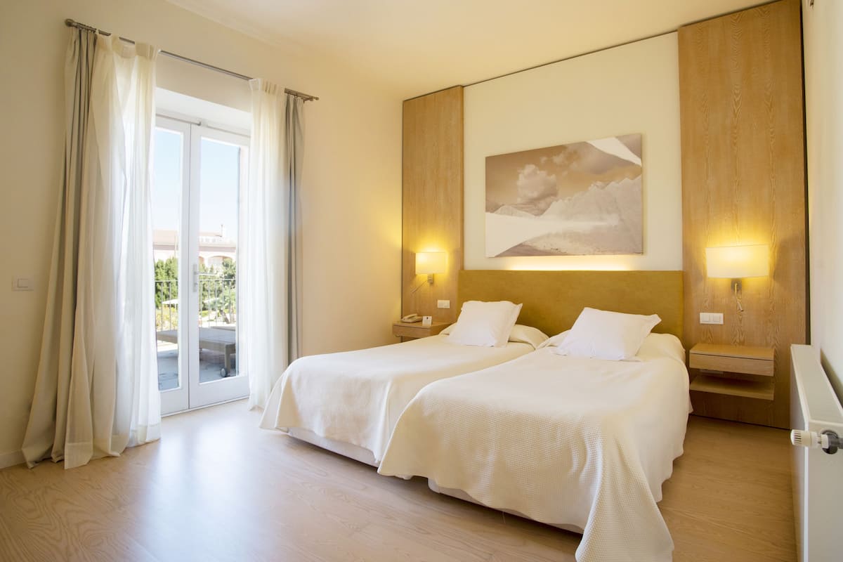 A hotel room with two beds, a large window with curtains, a painting on the wall, and a wooden nightstand with a lamp.