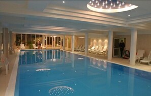 Une piscine intérieure à l’eau claire, entourée de chaises longues et dotée d’un plafond avec des spots encastrés.