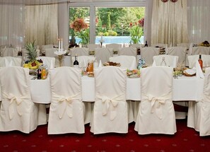 A banquet hall with round tables covered in white tablecloths, each with a centerpiece of fruit and candles. The room has a red carpet and large windows with a view of greenery and a pool. - Ambasador Chojny Hotel Lodz (Lodz)