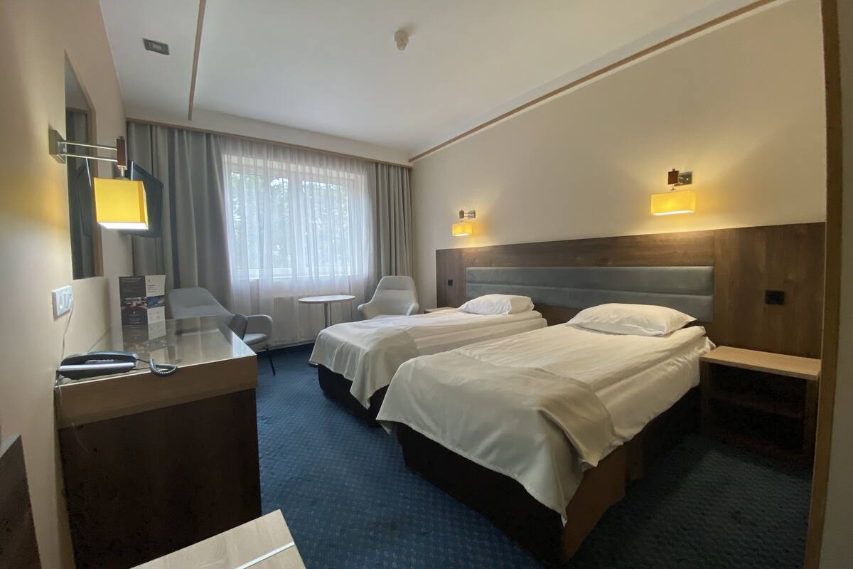 Habitación de hotel con dos camas, un escritorio con teléfono, una silla, una mesita y una ventana con cortinas.