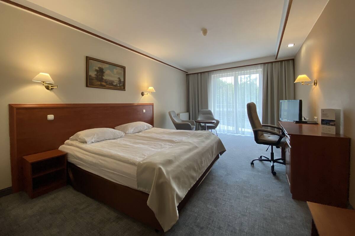 Habitación de hotel con una cama grande, un escritorio con silla, un televisor y un ventanal con cortinas.