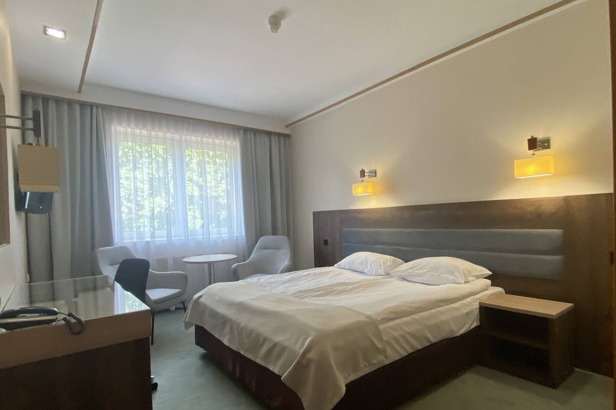 Habitación de hotel con una cama grande, un escritorio con silla, una mesita pequeña y un sofá. Hay dos luces fijadas a la pared y una ventana con cortinas.