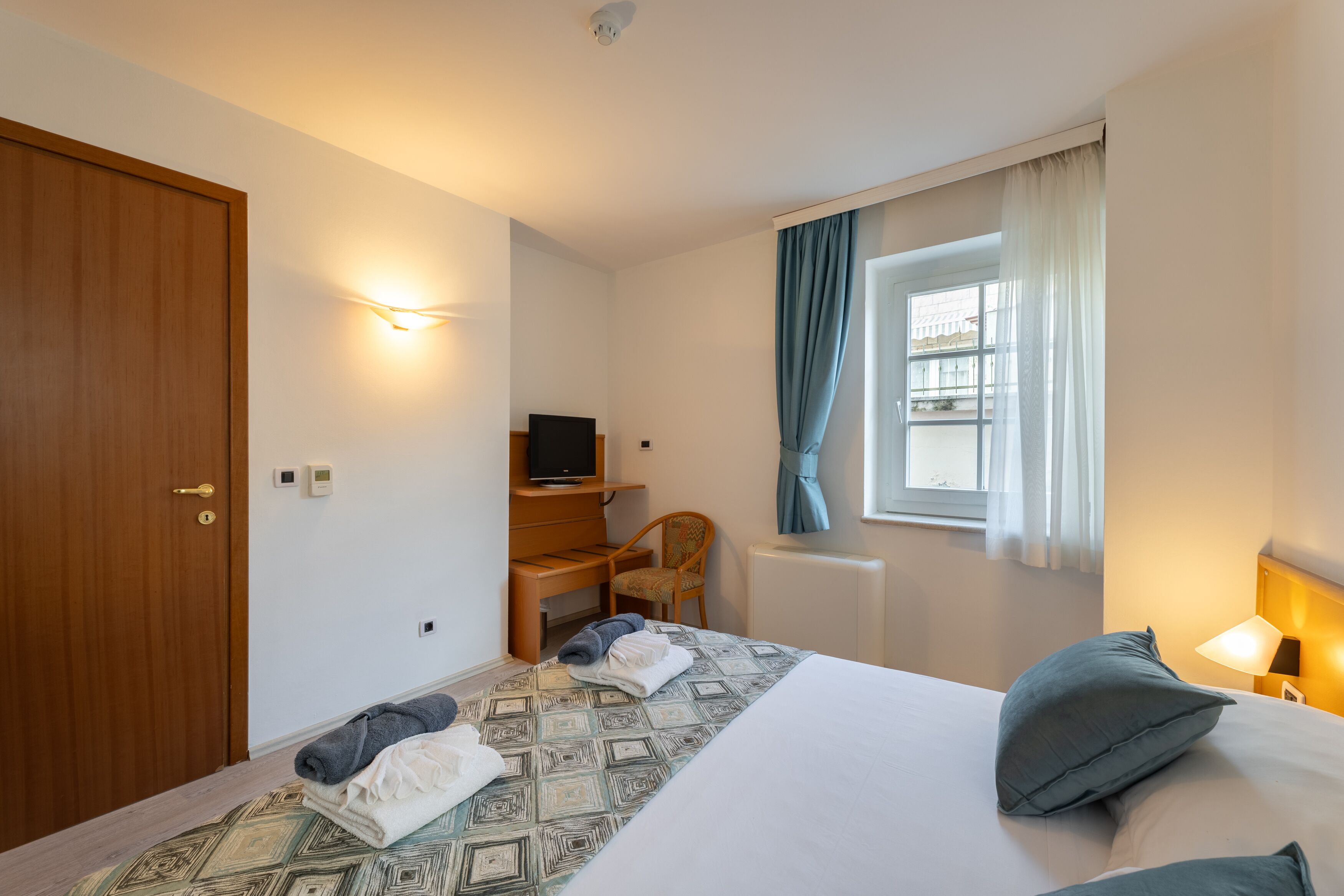 Una habitación de hotel con cama, escritorio con computadora, silla, ventana con cortinas y puerta.