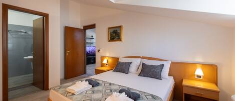 A hotel room with a bed, two bedside tables with lamps, a wooden headboard, a painting on the wall, and a bathroom visible through an open door.
