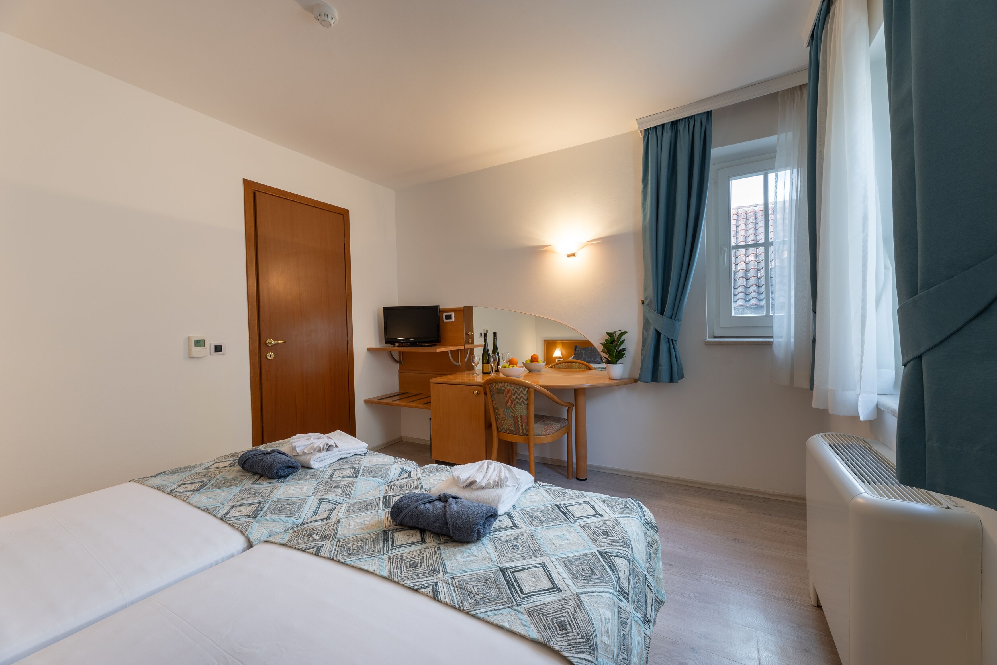 A hotel room with a bed, a desk, a chair, a TV, a window with curtains, and a heater.