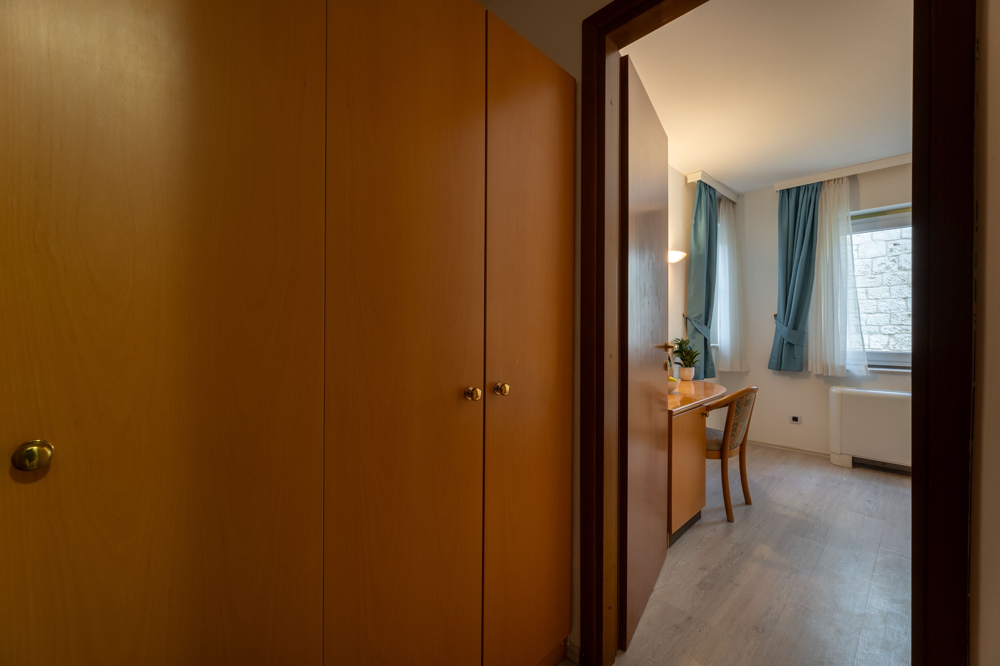 A room with a wooden wardrobe, a dining table with chairs, a potted plant, and a window with curtains.