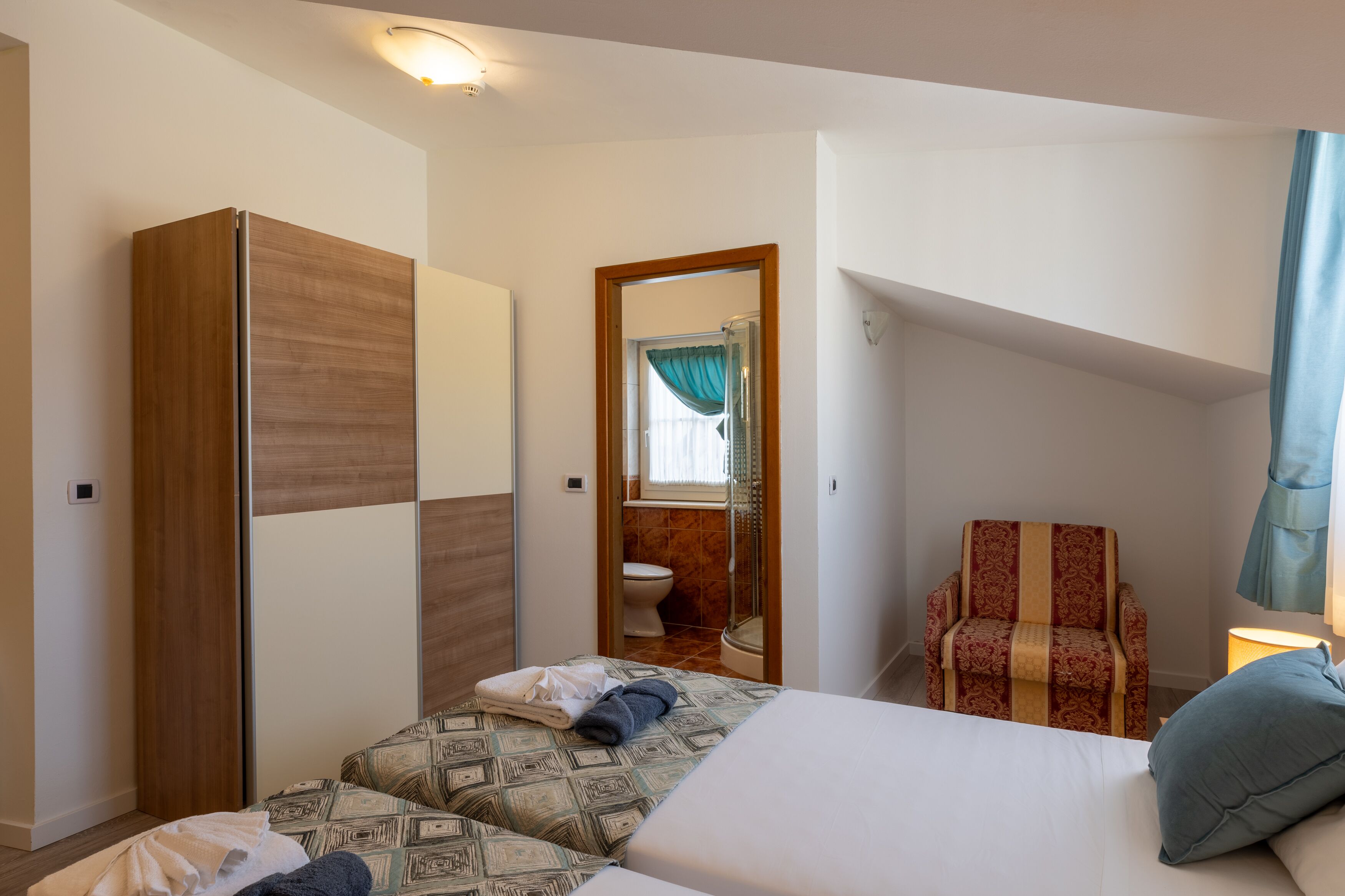 Un dormitorio con una cama, dos mesitas de noche, un armario, una silla y un baño visible a través de una puerta abierta.