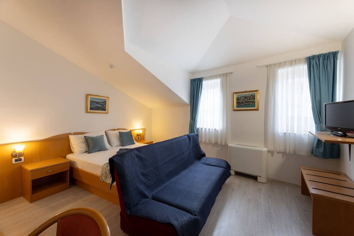 A hotel room with a bed, a sofa, a TV, a desk, and two paintings on the walls.
