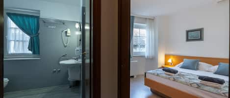 A hotel room with a bathroom, a bed with a headboard, a bedside table with a lamp, and a window with curtains.